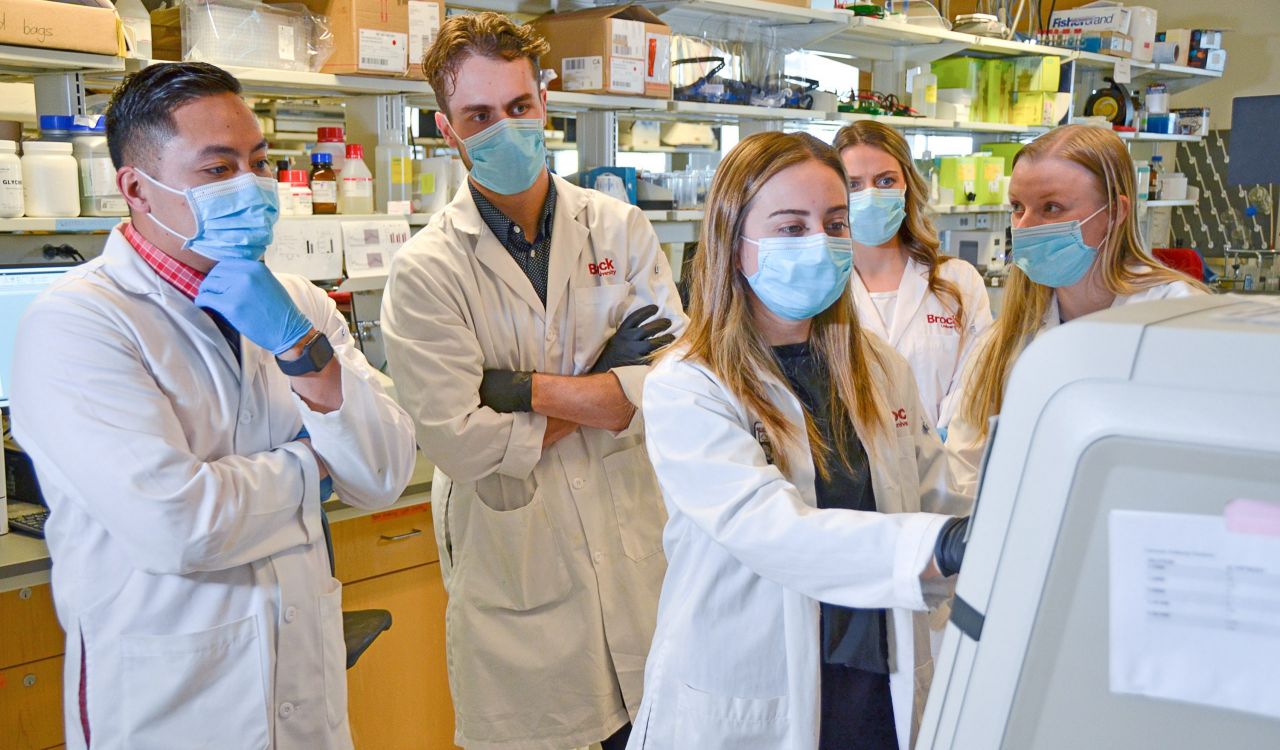 A group of masked researchers stand around a machine in a lab, looking at the screen. From left: Brock University Associate Professor of Kinesiology and Canada Research Chair in Tissue Plasticity and Remodelling Val Fajardo, master’s student Ryan Baranowski, PhD student Jessica Braun, master’s student Briana Hockey and master’s student Emily Copeland.