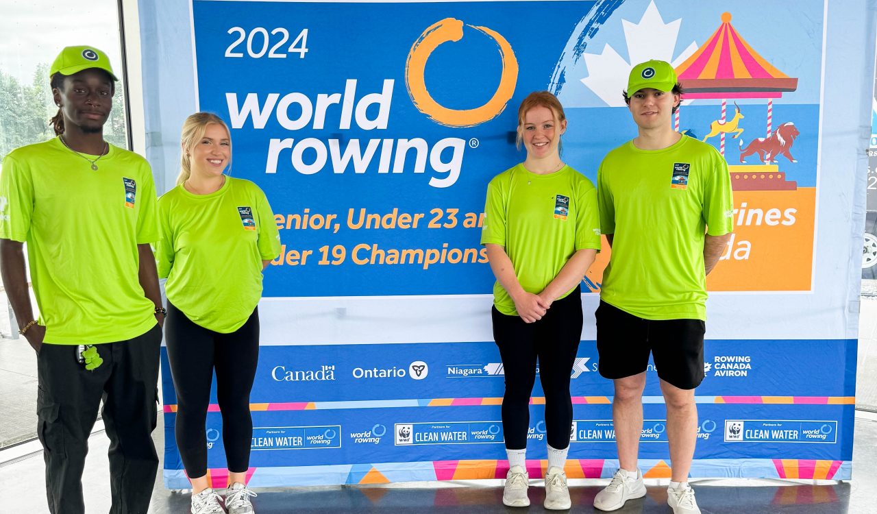 Four Brock University Sport Management students stand in front of a 2024 World Rowing Championships sign .