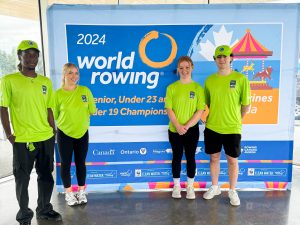 Four Brock University Sport Management students stand in front of a 2024 World Rowing Championships sign .