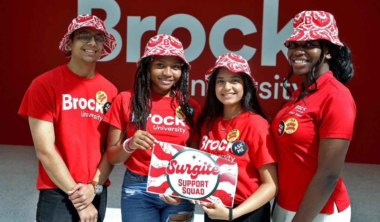 Four University students wearing red Brock shirts and tie-dye bucket hats stand in front of a red Brock University sign while holding a Surgite Support Squad Sign.