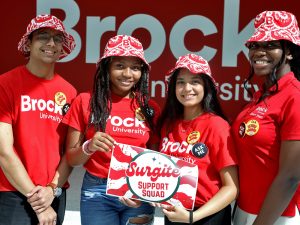 Four University students wearing red Brock shirts and tie-dye bucket hats stand in front of a red Brock University sign while holding a Surgite Support Squad Sign.