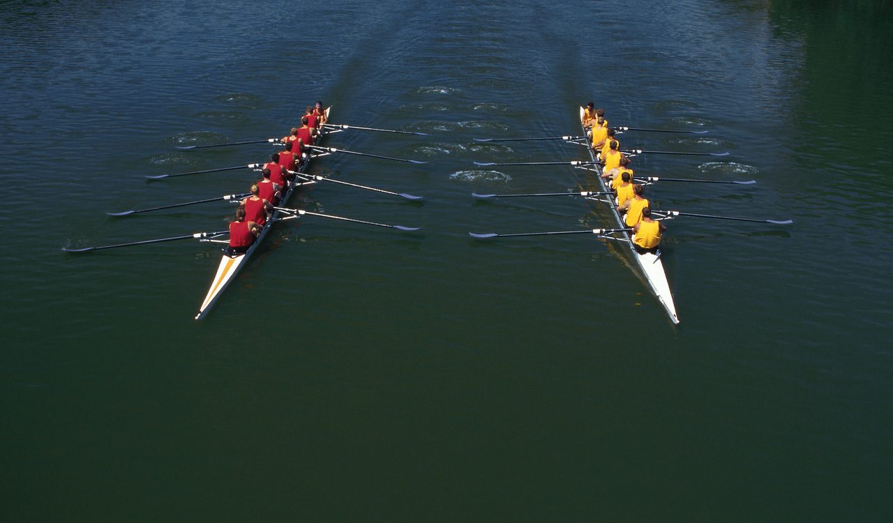 Aerial image of two teams of rowers floating on boats in water side by side and wearing yellow and red shirts.