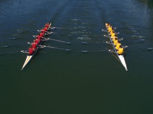 Aerial image of two teams of rowers floating on boats in water side by side and wearing yellow and red shirts.