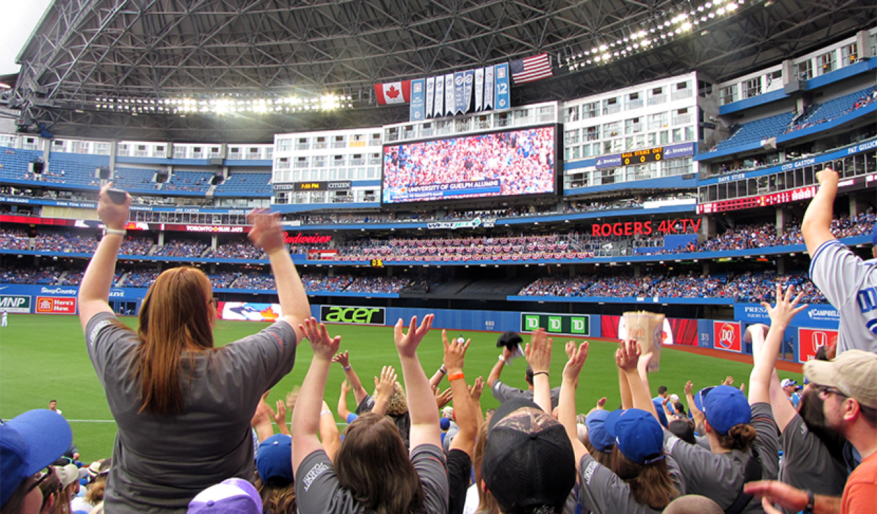 People, seen from behind, raise their arms while cheering for a baseball team in a stadium.