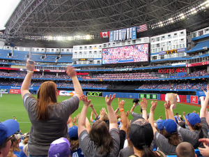 People, seen from behind, raise their arms while cheering for a baseball team in a stadium.