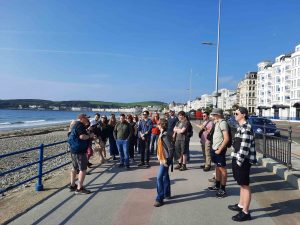 A group of people wearing backpacks and travel clothes stand on a promenade on a seashore against a brilliant blue sky.