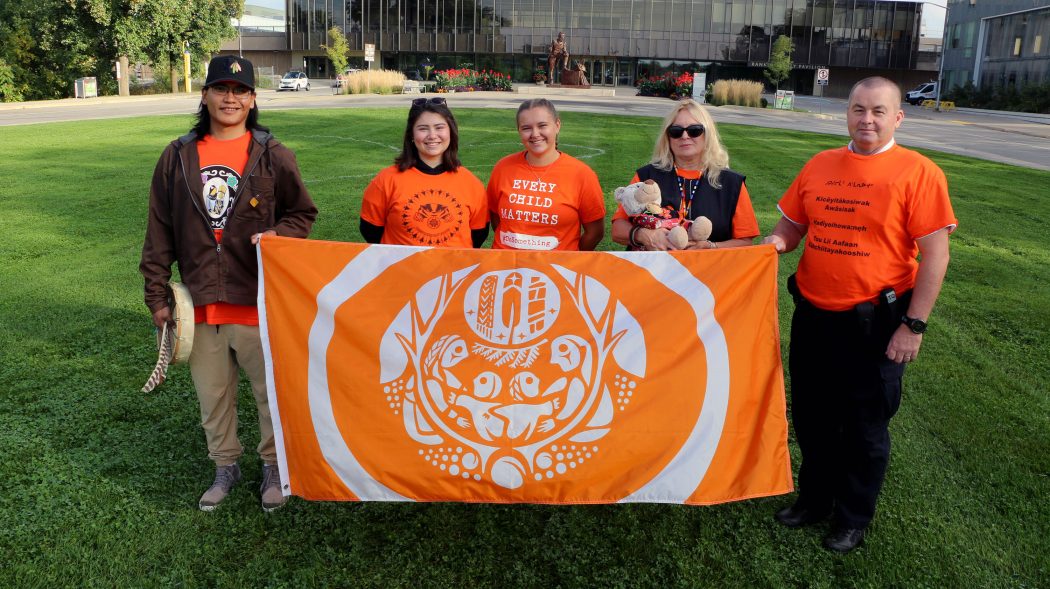 Five people stand together outside holding a bright orange and white flag, known as the Survivors' Flag.