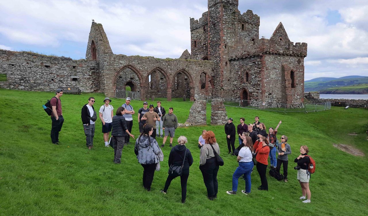 A group of university students on a picturesque island top with vibrant green grass in front of a historic stone castle site.