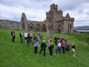 A group of university students on a picturesque island top with vibrant green grass in front of a historic stone castle site.