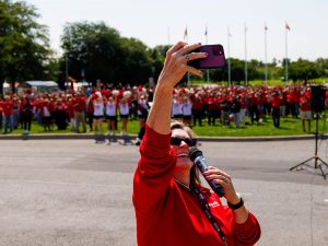 Brock University President and Vice-Chancellor Lesley Rigg takes a selfie with hundreds of people in the background while standing outside on a field.