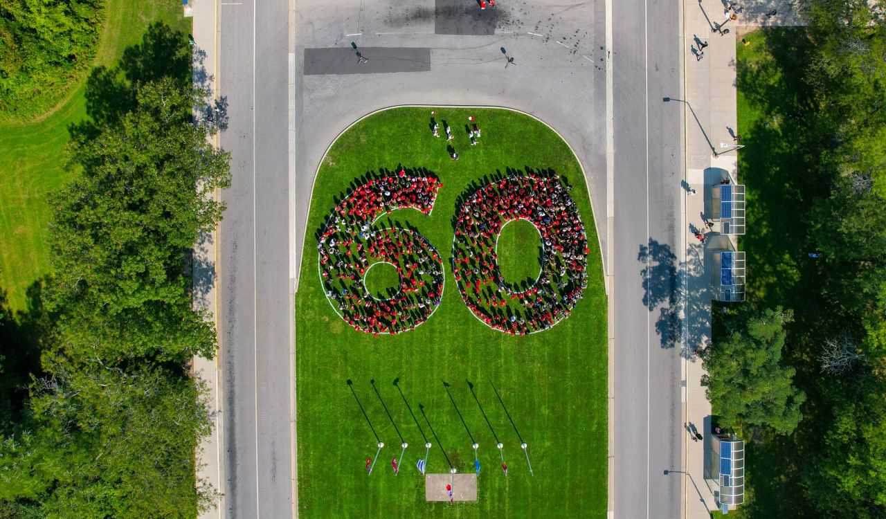 Hundreds of people wearing red shirts form a large 60 on the green lawn of a university. They are pictured from the sky.