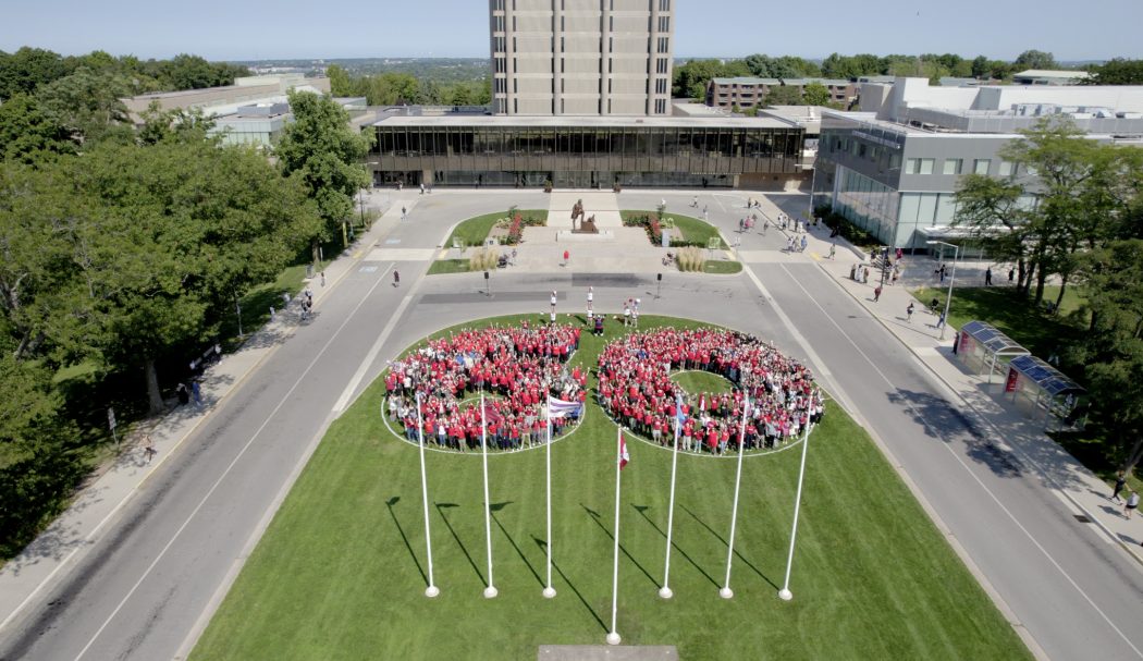 Hundreds of people wearing red shirts form a large 60 on the green lawn of a university. They are pictured from the sky.