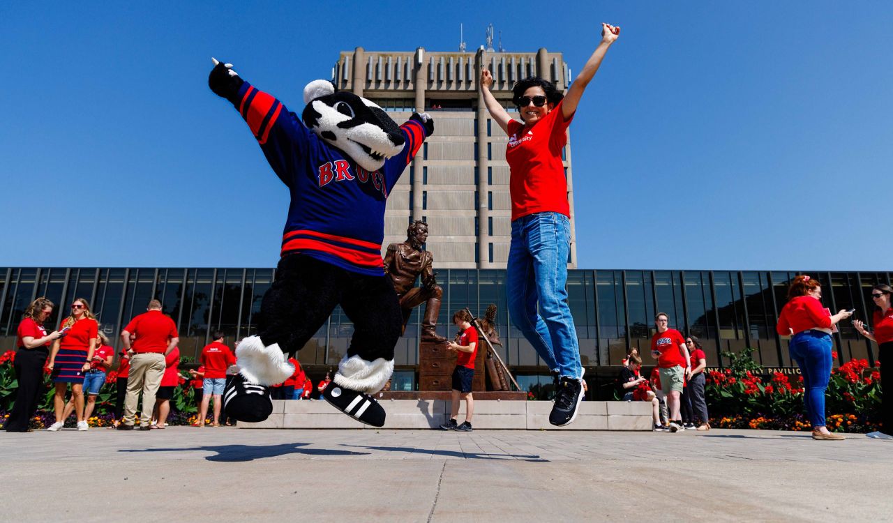 Boomer the Badger and a person in a red Brock shirt jump in the air in front of the iconic Schmon Tower.