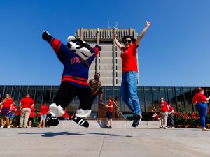 Boomer the Badger and a person in a red Brock shirt jump in the air in front of the iconic Schmon Tower.