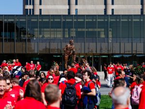 People in red shirts stand on a green field in front of Brock University's Rankin Family Pavilion and Schmon Tower.