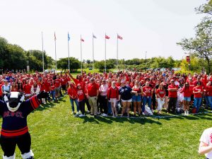 People in red shirts stand on a green field in front at Brock University. Brock's mascot, Boomer the Badger, stands in the foreground.