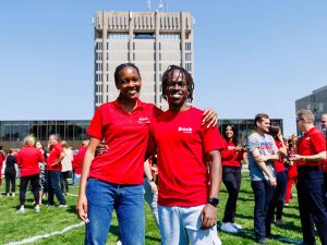 People in red shirts stand on a green field in front of Brock University's Rankin Family Pavilion and Schmon Tower.