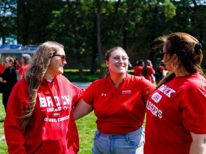 Three people in red Brock University shirts stand together looking at one another while outside on a green field.