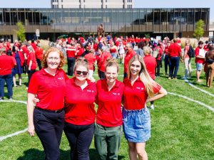 People in red shirts stand on a green field in front of Brock University's Rankin Family Pavilion and Schmon Tower.