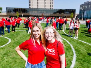 People in red shirts stand on a green field in front of Brock University's Rankin Family Pavilion and Schmon Tower.