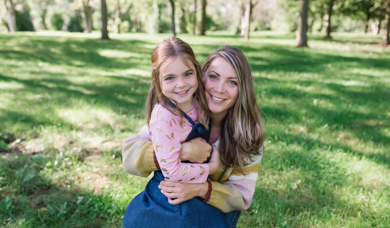 A young girl and her mother smile and hug for a photo in a park.