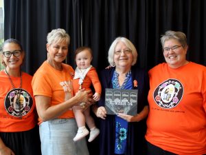 Four women and a baby stand together. Three women and the baby are dressed in orange in recognition of Orange Shirt Day.