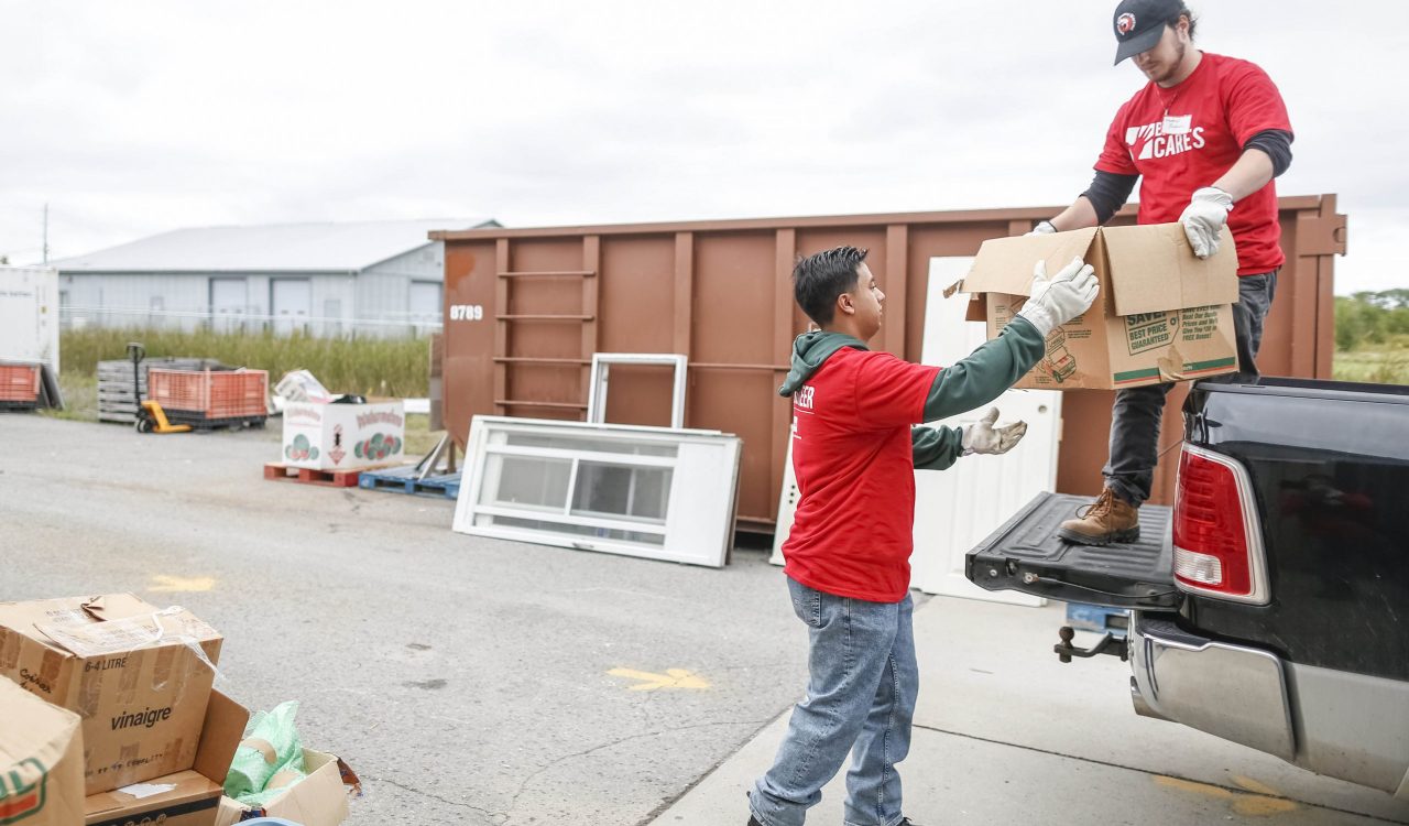 Two university students in red shirts load boxes onto the back of a truck in a parking lot during a volunteer event.