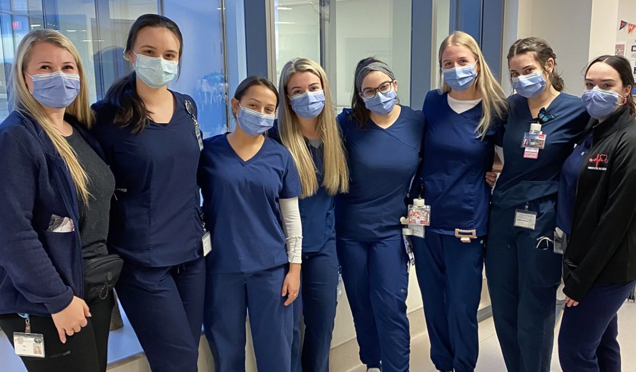 Eight Brock University Nursing students wearing navy-coloured scrubs and face masks stand next to each other in a line.