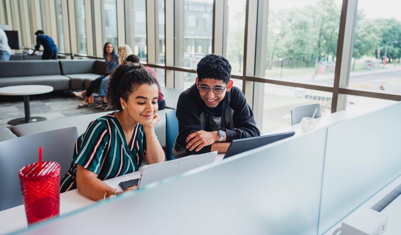 Two university students work at a desk in front of a wall of windows.