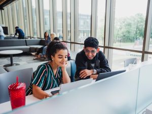 Two university students work at a desk in front of a wall of windows.