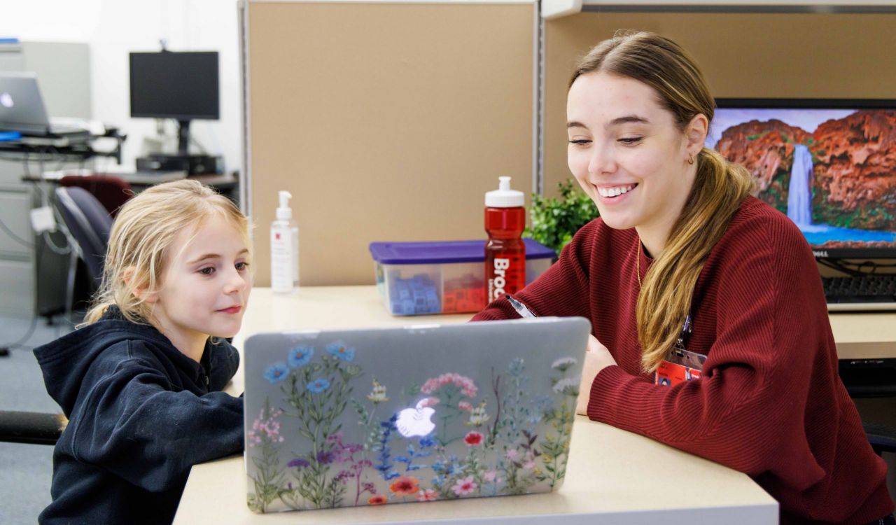 A young woman sits at a table across from a school-aged child while both look at a laptop that is decorated with colourful flower stickers. Both people are smiling warmly in the well-lit, bright room.