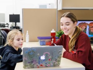 A young woman sits at a table across from a school-aged child while both look at a laptop that is decorated with colourful flower stickers. Both people are smiling warmly in the well-lit, bright room.