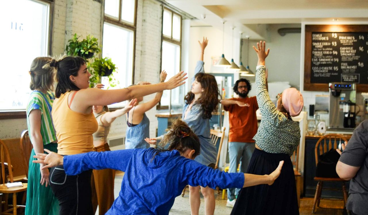 A group of people stand in a room with white brick walls and a coffee counter in the background, moving together with arms in the air and people in various positions invoking a sense of improvisation.