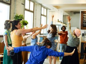 A group of people stand in a room with white brick walls and a coffee counter in the background, moving together with arms in the air and people in various positions invoking a sense of improvisation.