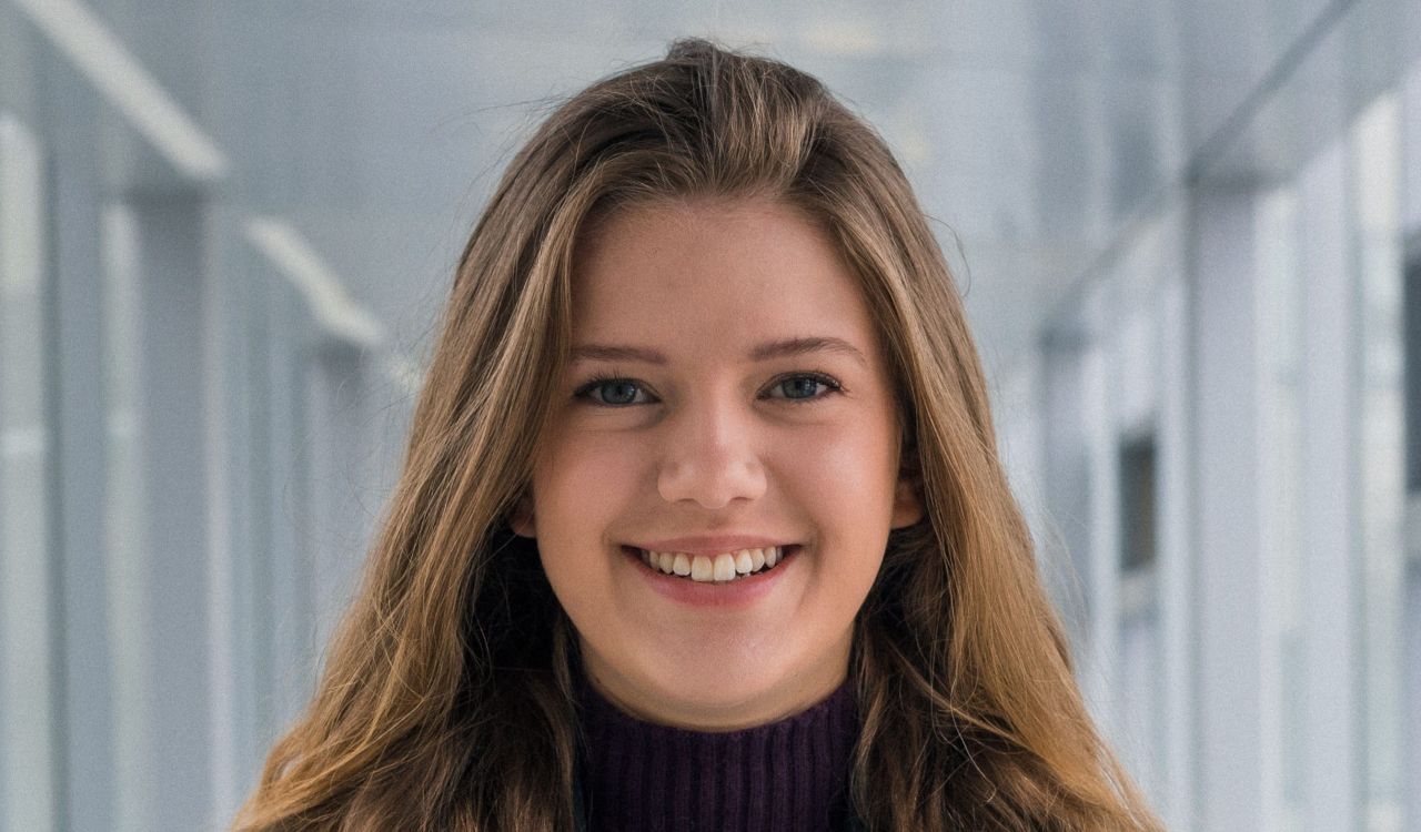 Carleigh Charlton is pictured in front a long hallway wearing a blue blazer and purple shirt.