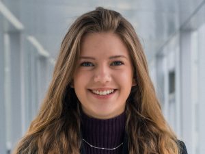 Carleigh Charlton is pictured in front a long hallway wearing a blue blazer and purple shirt.