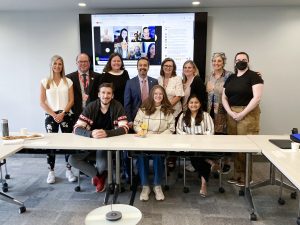 A group of people at Brock University stand together at a board room table in front of a digital screen to celebrate an award recipient.