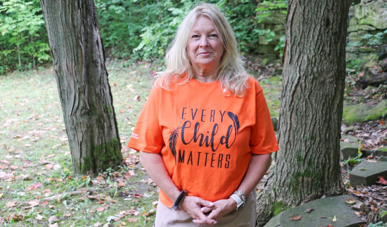 Cindy Biancaniello wears an orange shirt and stands in front of an outdoor rock wall surrounded by trees at Brock University.