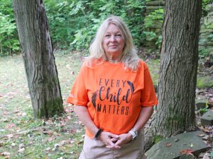 Cindy Biancaniello wears an orange shirt and stands in front of an outdoor rock wall surrounded by trees at Brock University.