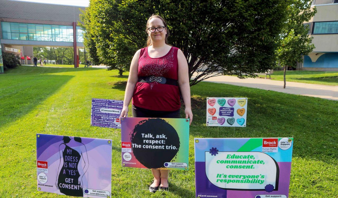 Katie Keays stands outside in the grass among a four lawn signs featuring text and graphics about consent-related issues.