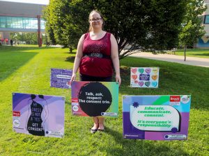 Katie Keays stands outside in the grass among a four lawn signs featuring text and graphics about consent-related issues.