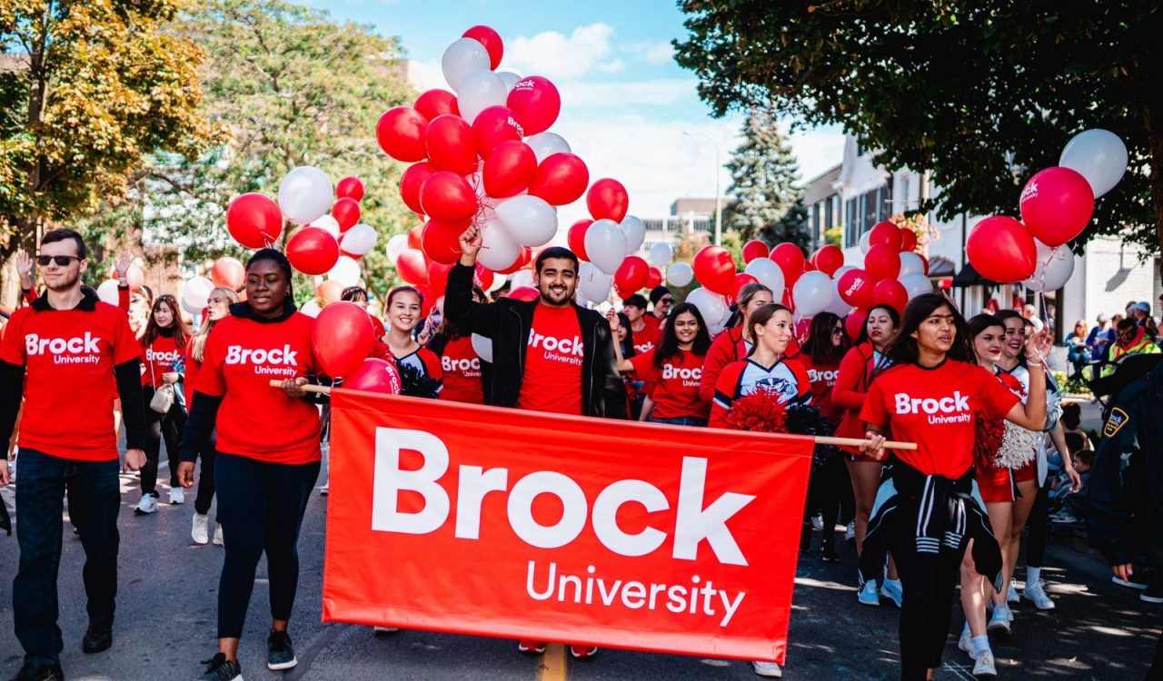 People in red shirts walk behind a Brock University banner while holding balloons at a parade.