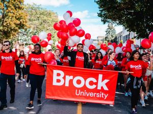 People in red shirts walk behind a Brock University banner while holding balloons at a parade.