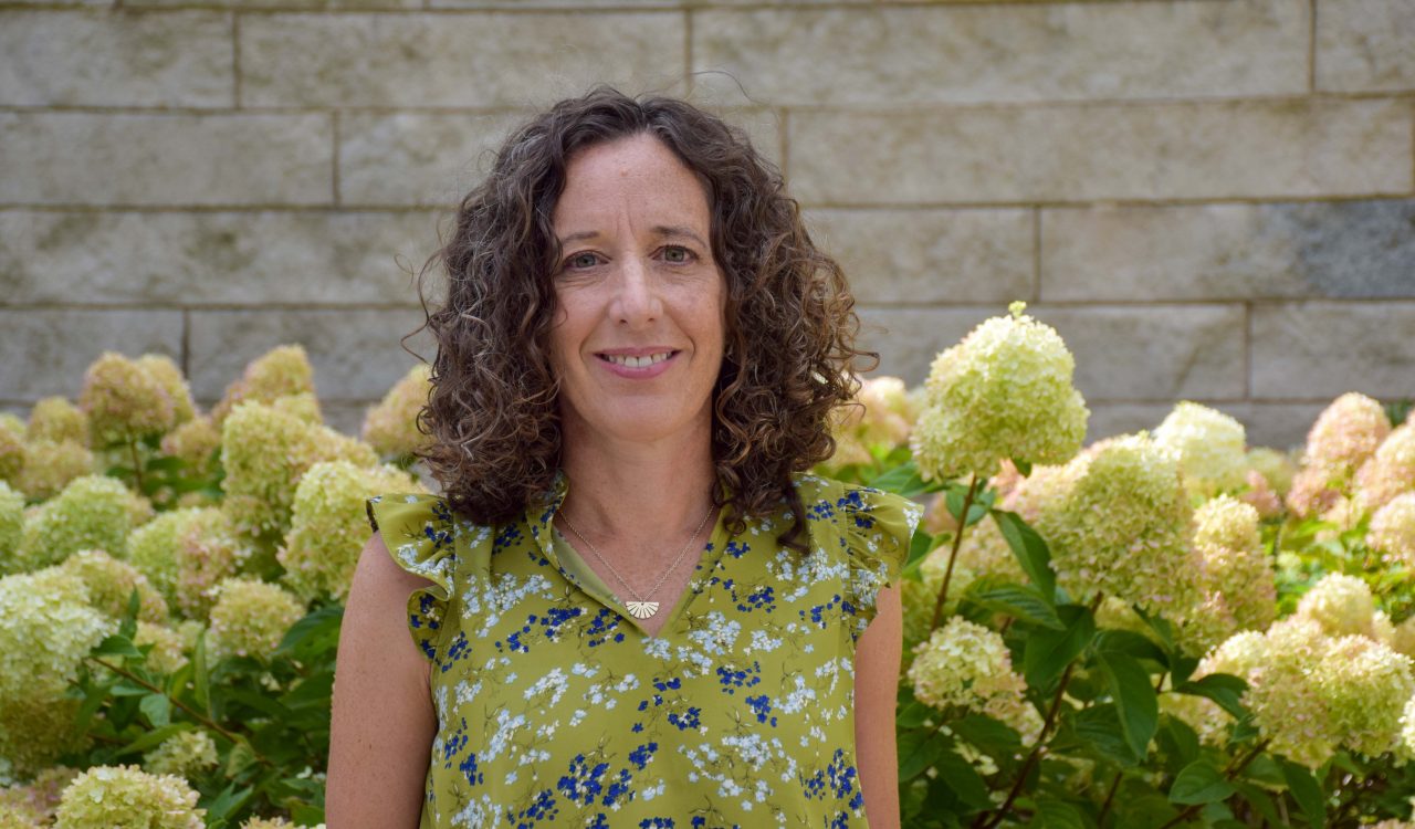 A woman in a floral shirt poses for a photo in front of blooming hydrangeas and stone wall.