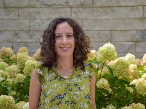 A woman in a floral shirt poses for a photo in front of blooming hydrangeas and stone wall.