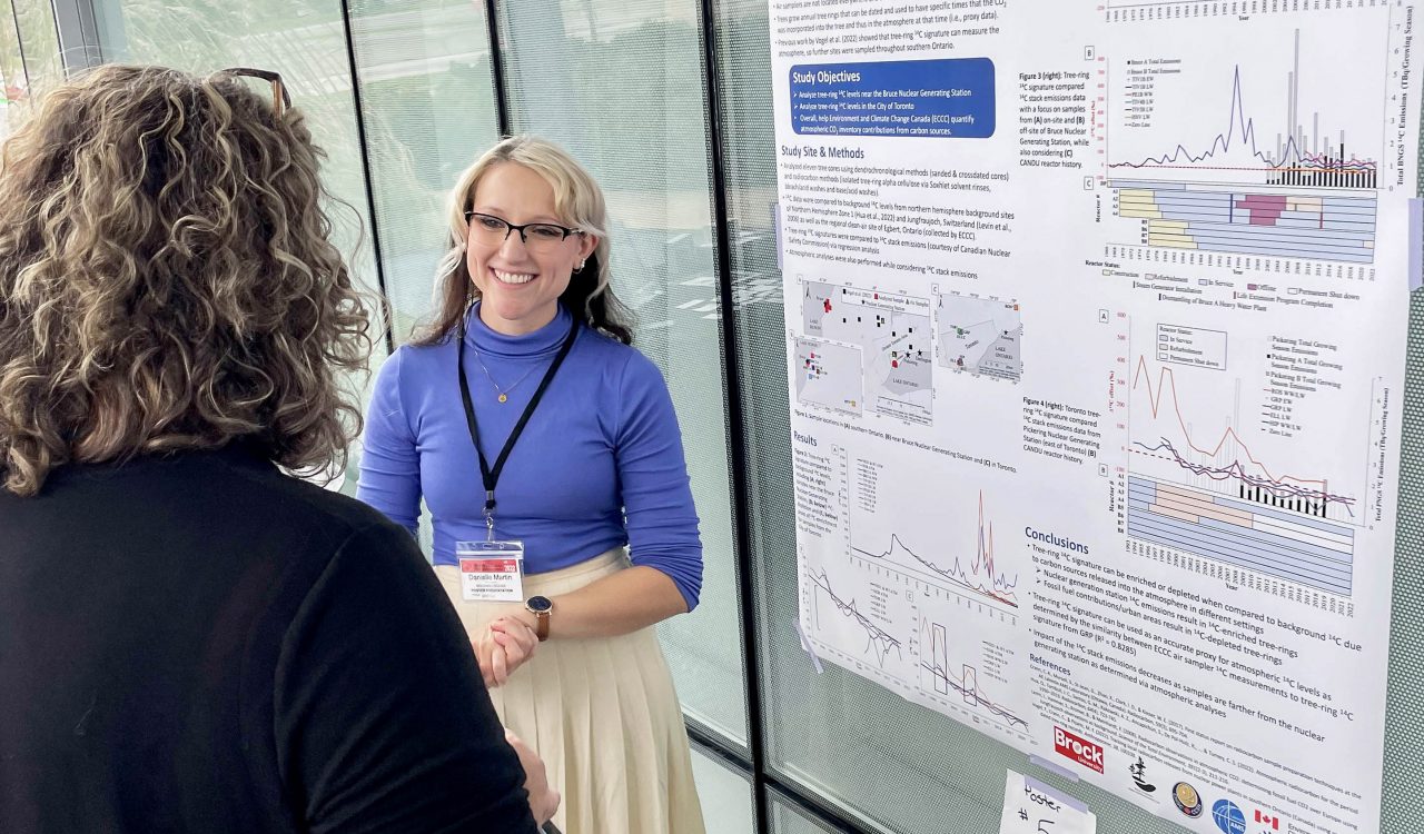 Brock University master’s student Danielle Martin smiles as she speaks with a judge about her research, which is displayed on a large poster behind her entitled, “Estimating atmospheric 14C using tree rings to monitor nuclear generating station emissions and fossil fuel contributions in southern Ontario, Canada.”