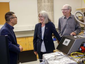 Three people stand close to each other talking. Beside them is a table with scientific lasers used in research.