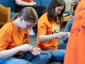 Two people in orange shirts sit cross-legged on the ground while making dream catchers.