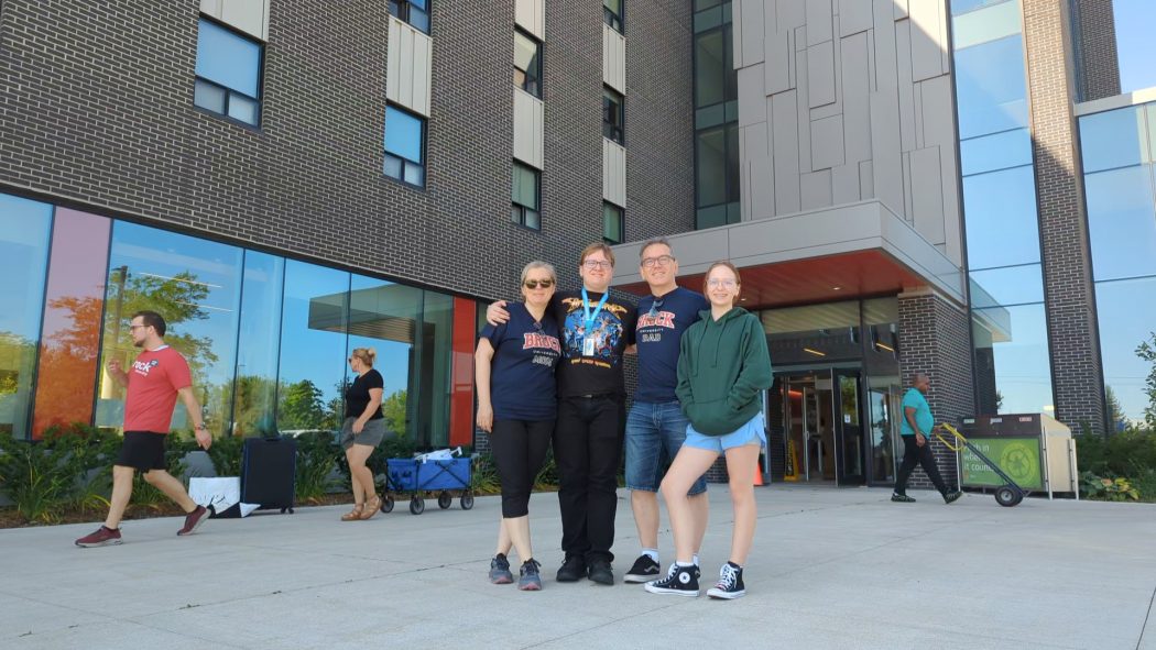 A family of four stands in front of a tall residence building as people with trolleys walk in the background.