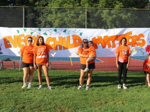 A group of eight adults and children, all in orange shirts, stand in front of a banner that reads: Every Child Matters.